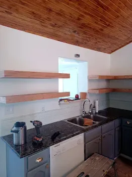Kitchen with gray cabinets, dark granite countertops, floating wood shelves, and a sloped wood-paneled ceiling.