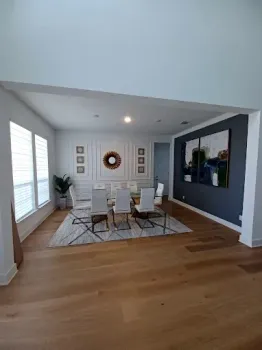 A bright dining room with hardwood floors, a white rug, six chairs, and a dark accent wall featuring large art.