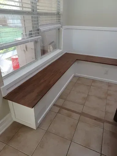 An L-shaped built-in bench with a dark wood seat and white base positioned under a window in a tiled room.