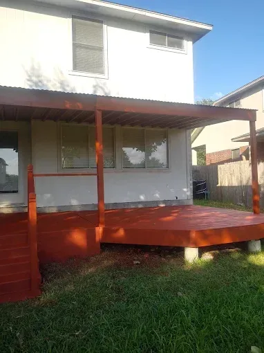 A newly stained, reddish-brown wooden deck and covered porch attached to the back of a white two-story house.