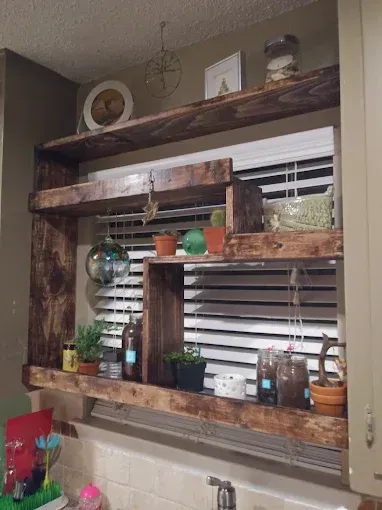 Dark wood rustic shelves mounted over a kitchen window filled with various plants, decor items, and glass jars.