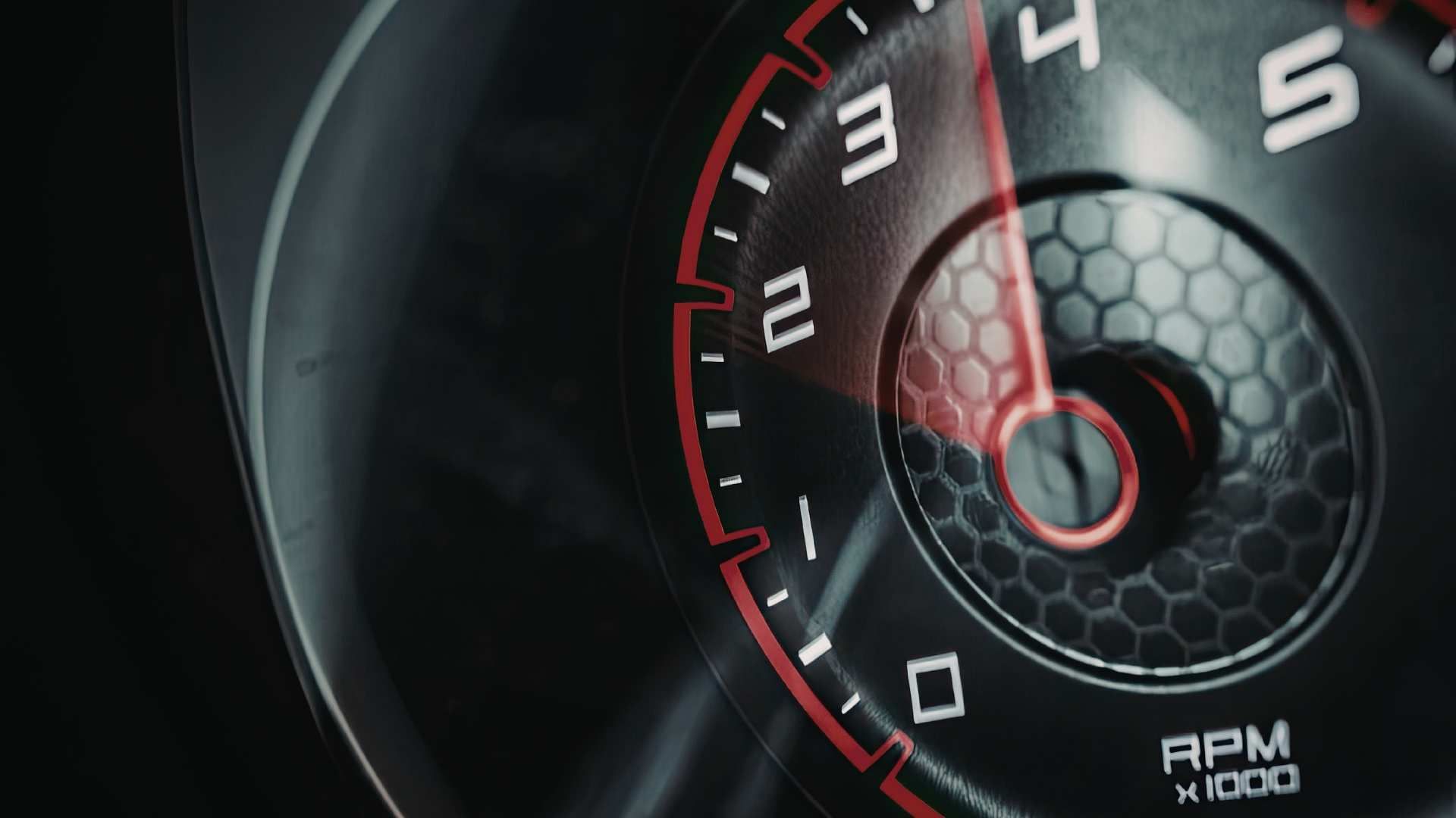 Close-up of a dark car tachometer gauge with a red needle pointing near 4,000 RPM on a hexagonal patterned background.