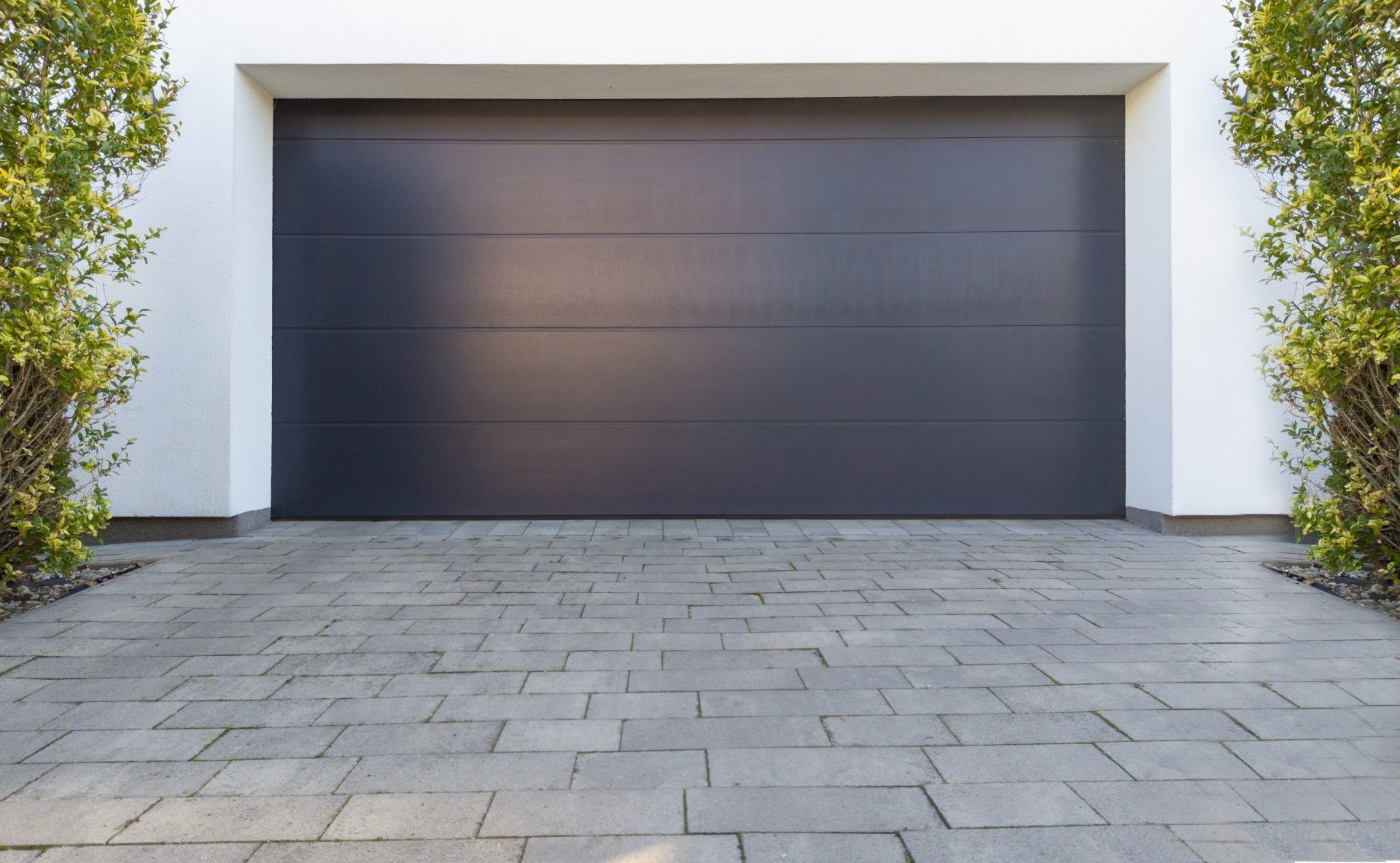 A modern dark gray garage door centered between two green shrubs, with a gray stone paved driveway in the foreground.
