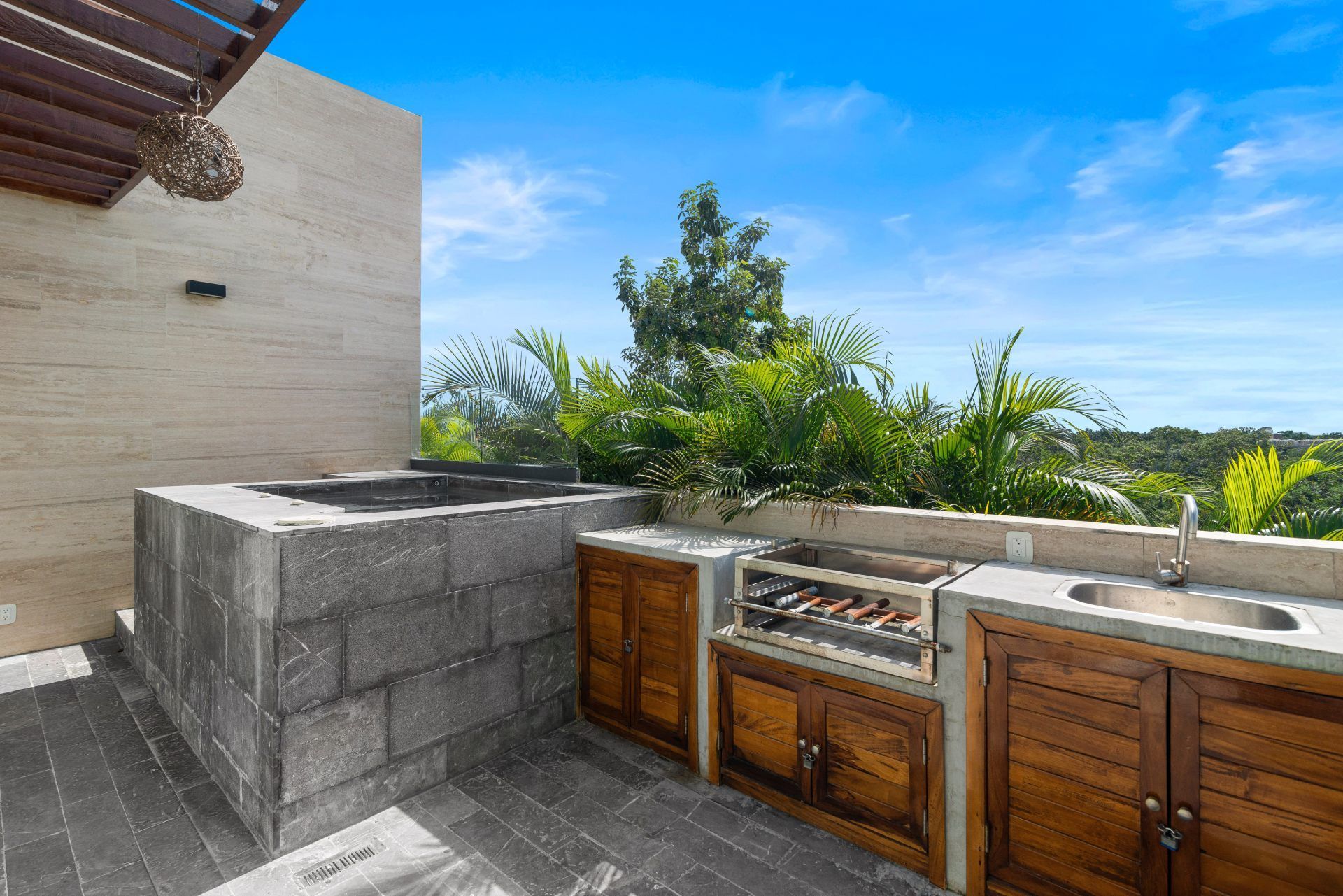 Outdoor kitchen and stone hot tub on a tiled patio under a blue sky, featuring wooden cabinets, a grill, and a sink.