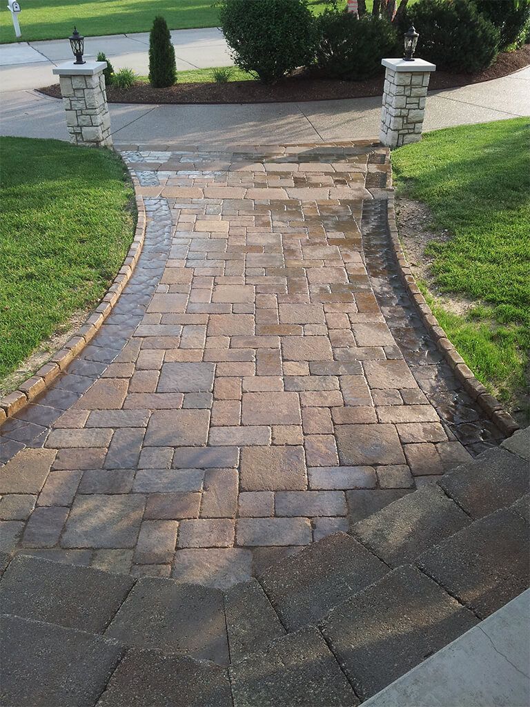 Brick paver walkway leading from a home, bordered by green grass and two stone columns.