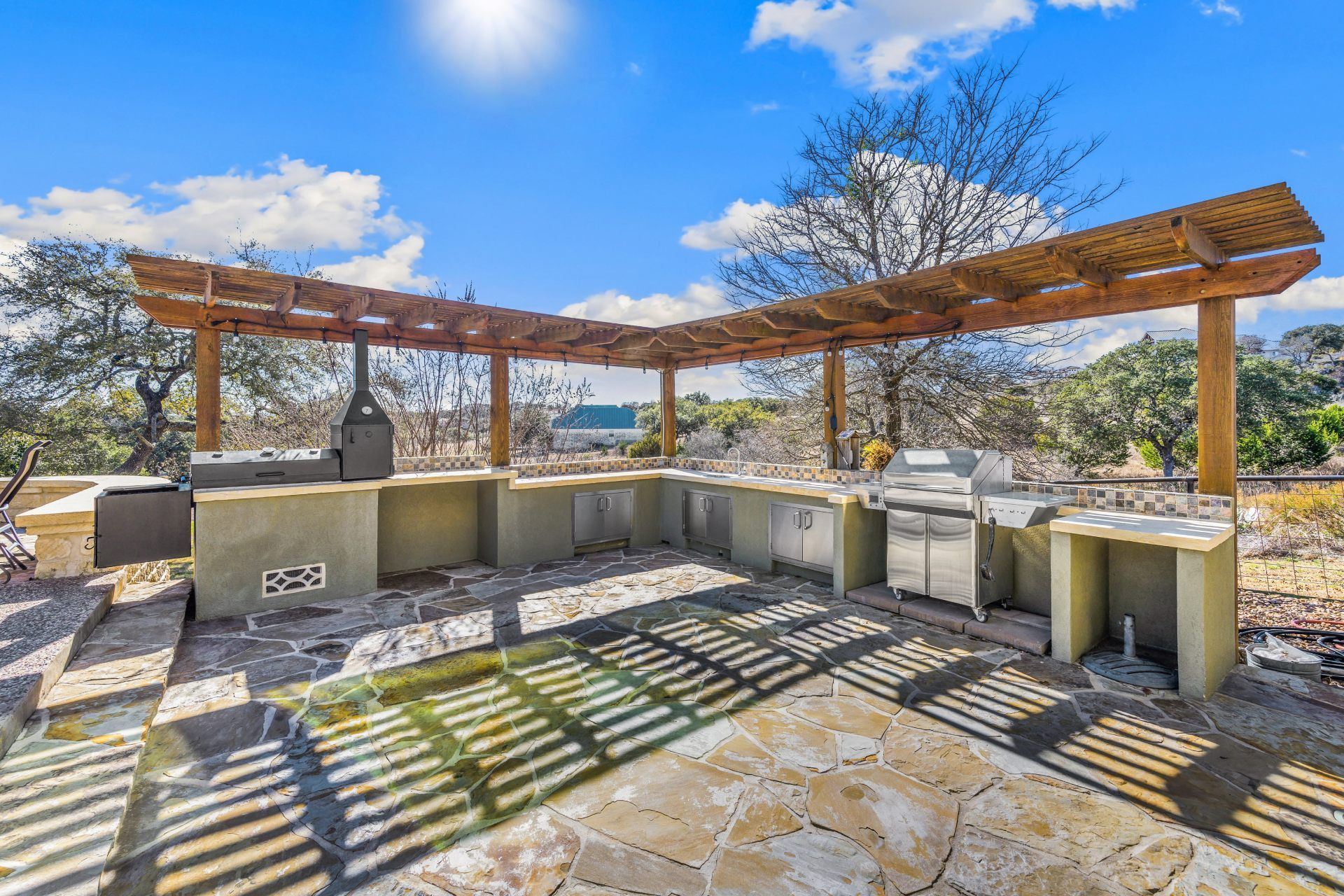 Outdoor kitchen with pergola, stone patio, grill, and counter space.