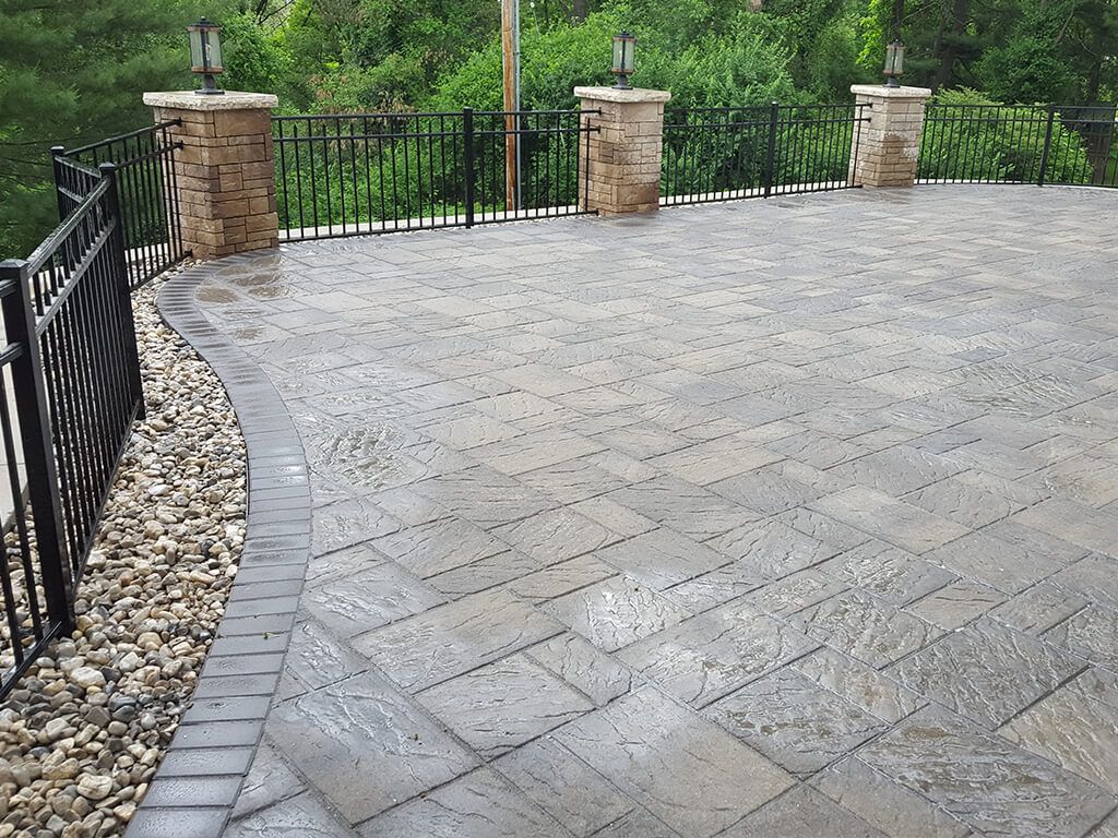 Concrete patio with black metal railing, stone columns, and surrounding greenery.