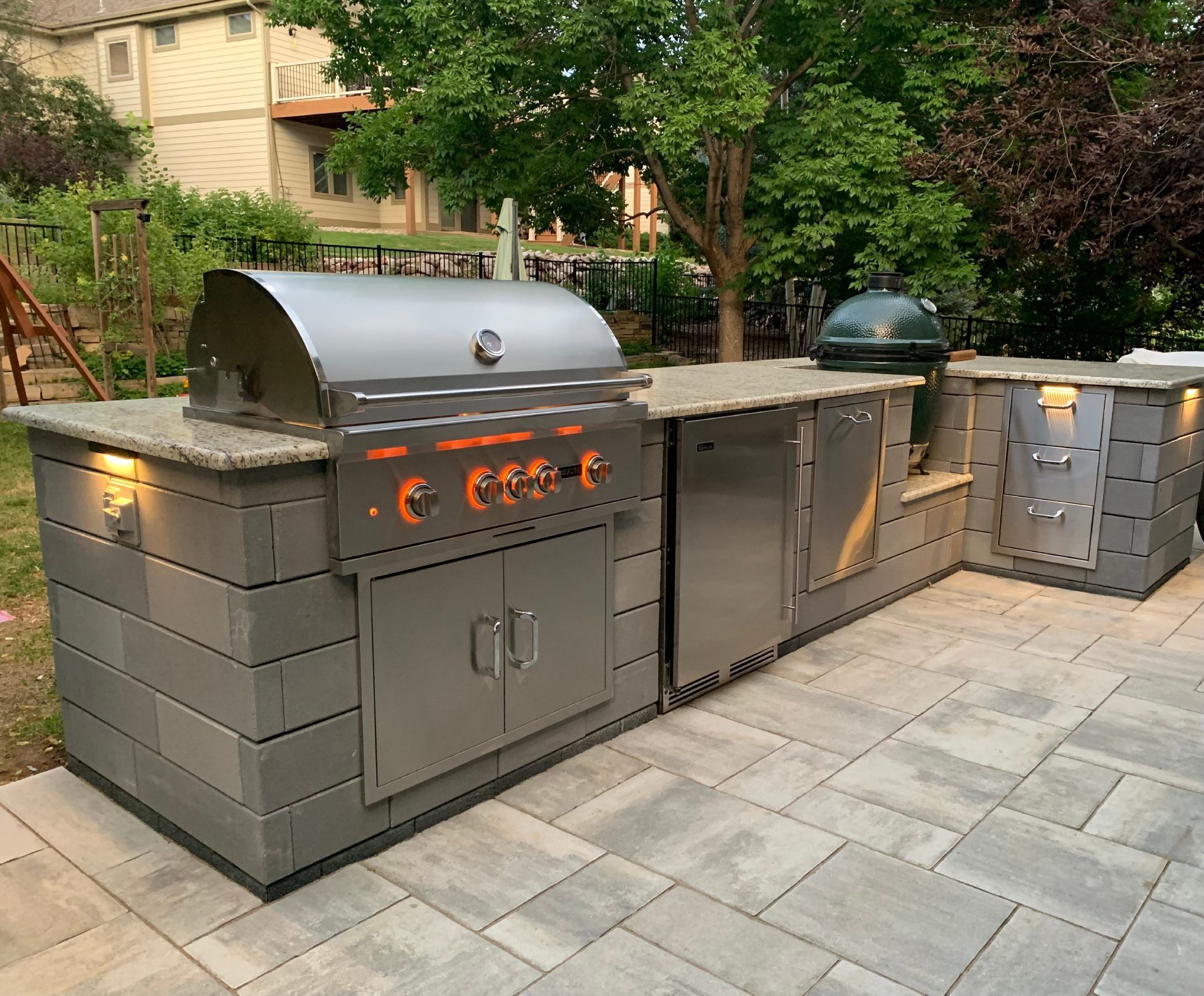 Outdoor grilling station with stainless steel appliances, stone countertop, gray brick, and a patio setting.