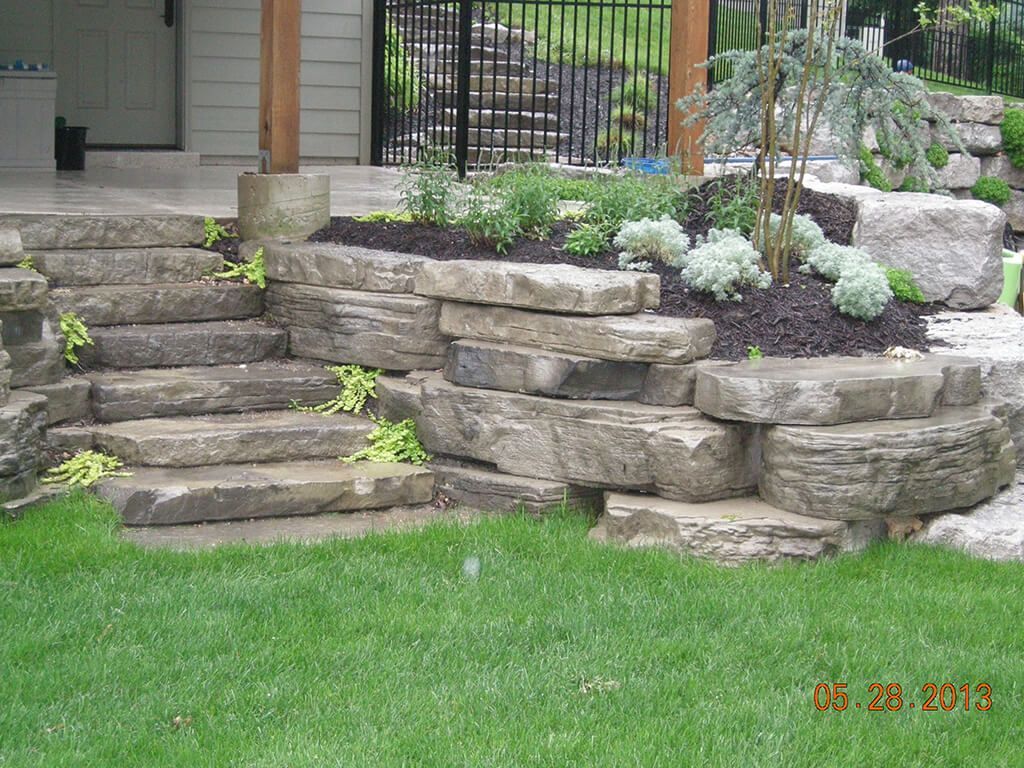 Stone steps leading up to a house entrance, with tiered garden beds and green grass.