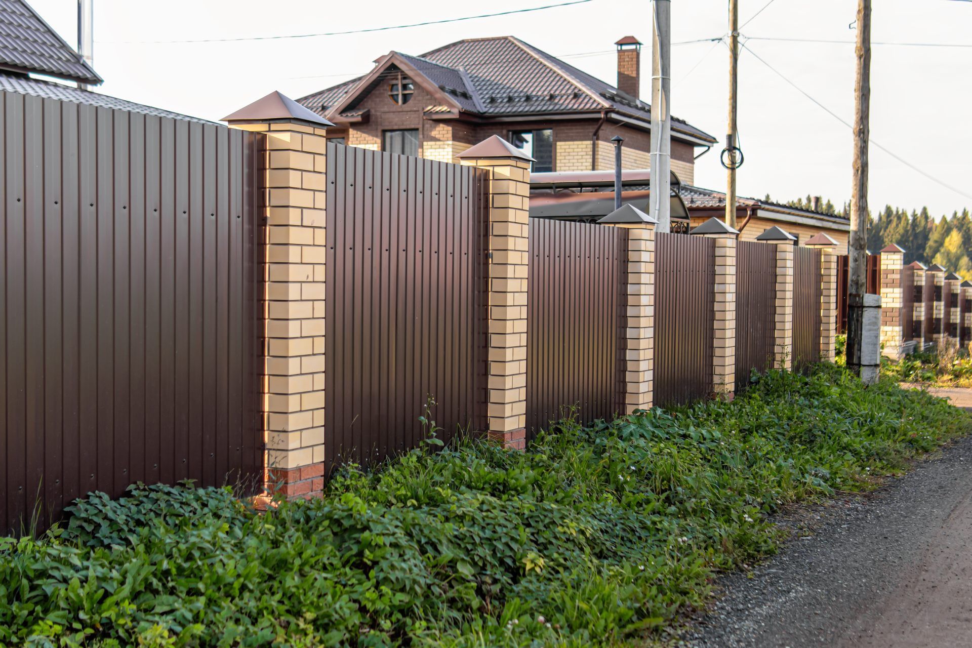 Brown metal fence with brick pillars bordering a house, green bushes in front.