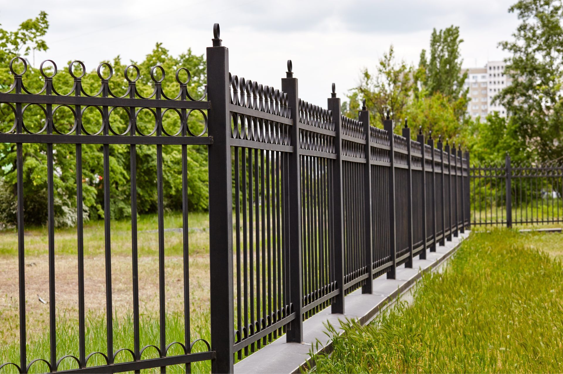 A black metal picket fence with circular decorative tops lining a grassy lawn under a cloudy sky.