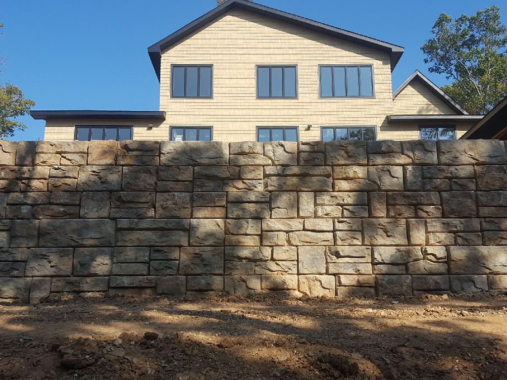 Stone retaining wall in front of a tan-colored house with dark window frames under a clear blue sky.