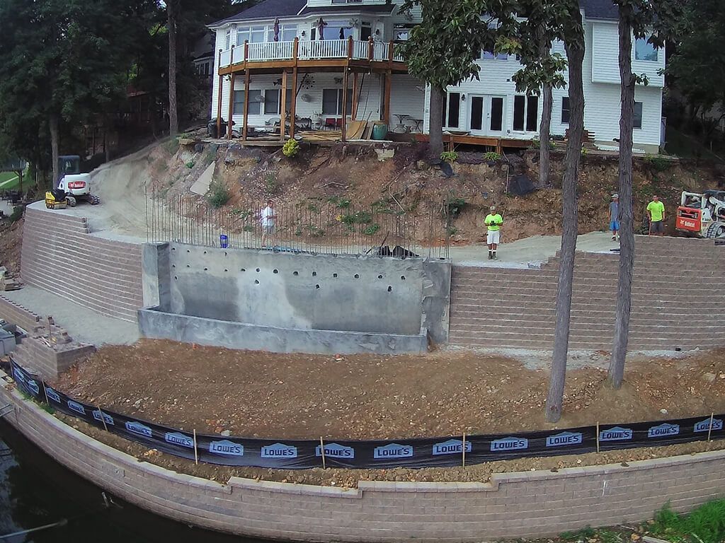 Construction site with retaining walls at lakeside house; workers and heavy machinery visible.