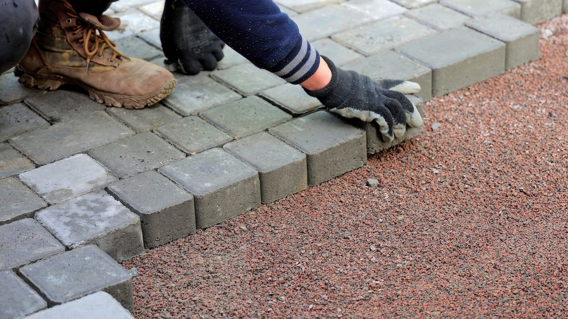 A worker's gloved hands lay grey rectangular paving stones onto a red gravel base.