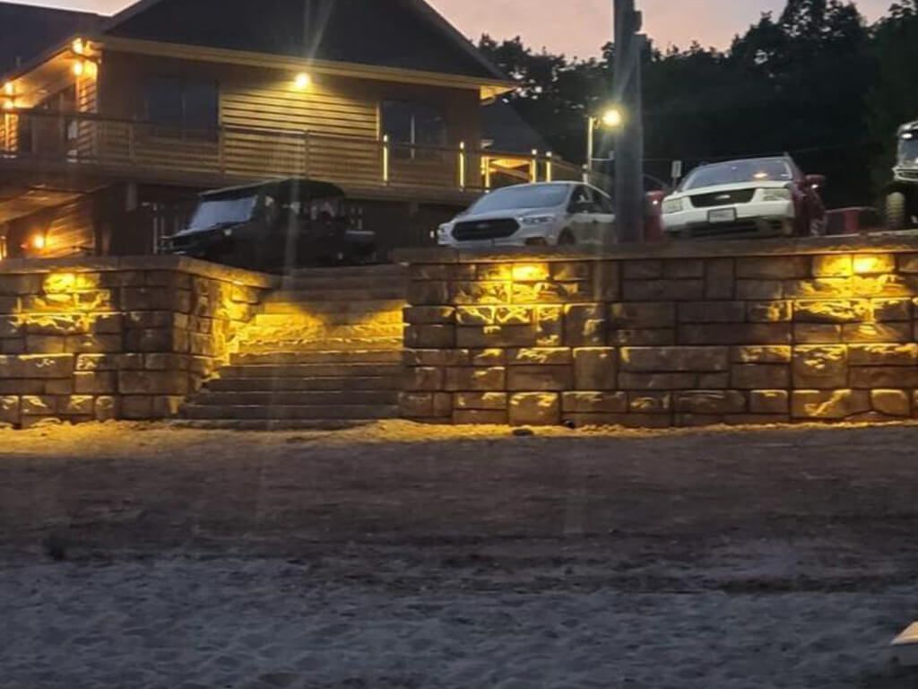 Lit stone retaining wall with steps leading to a house, cars parked above. Evening setting.