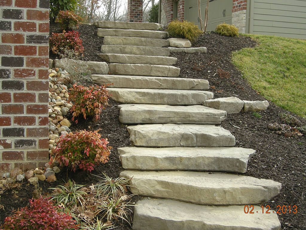 Stone steps leading uphill in a landscaped yard, flanked by brick wall and shrubs with mulch.