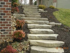 Stone steps leading uphill in a landscaped yard, flanked by brick wall and shrubs with mulch.