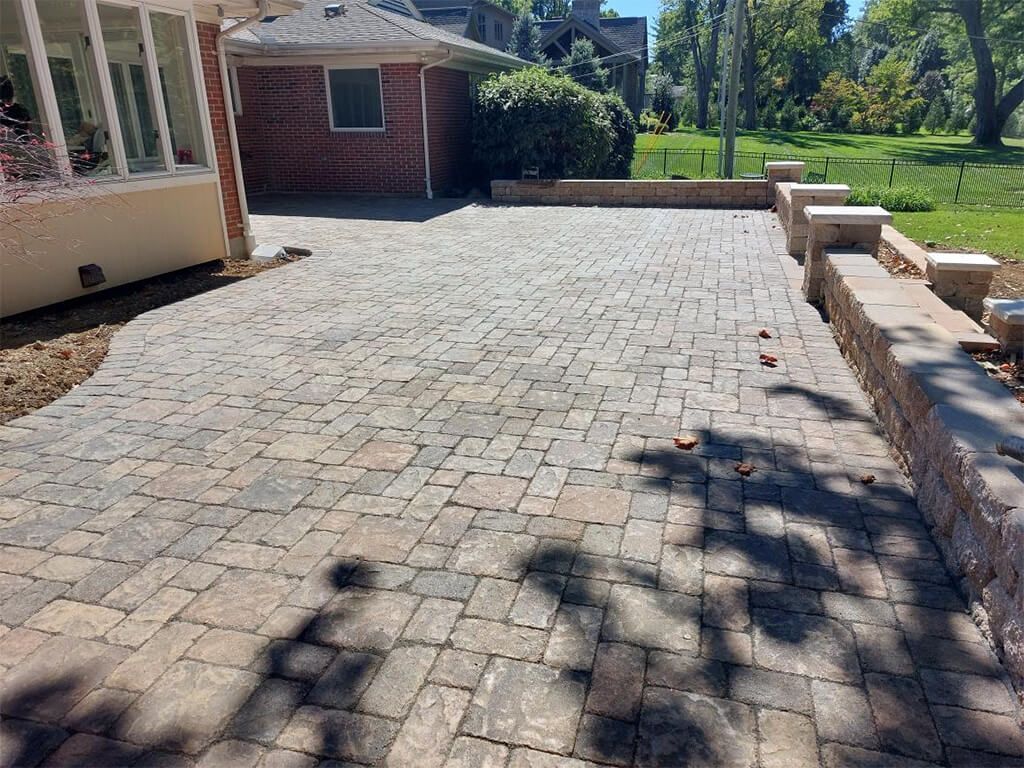 Stone-paved patio beside a house, with low brick seating wall and trees in bright sunlight.