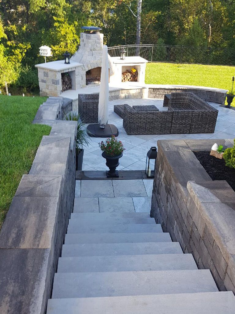 Stone steps lead up to a patio with a fireplace, seating area, and outdoor kitchen. Green lawn surrounds the space.