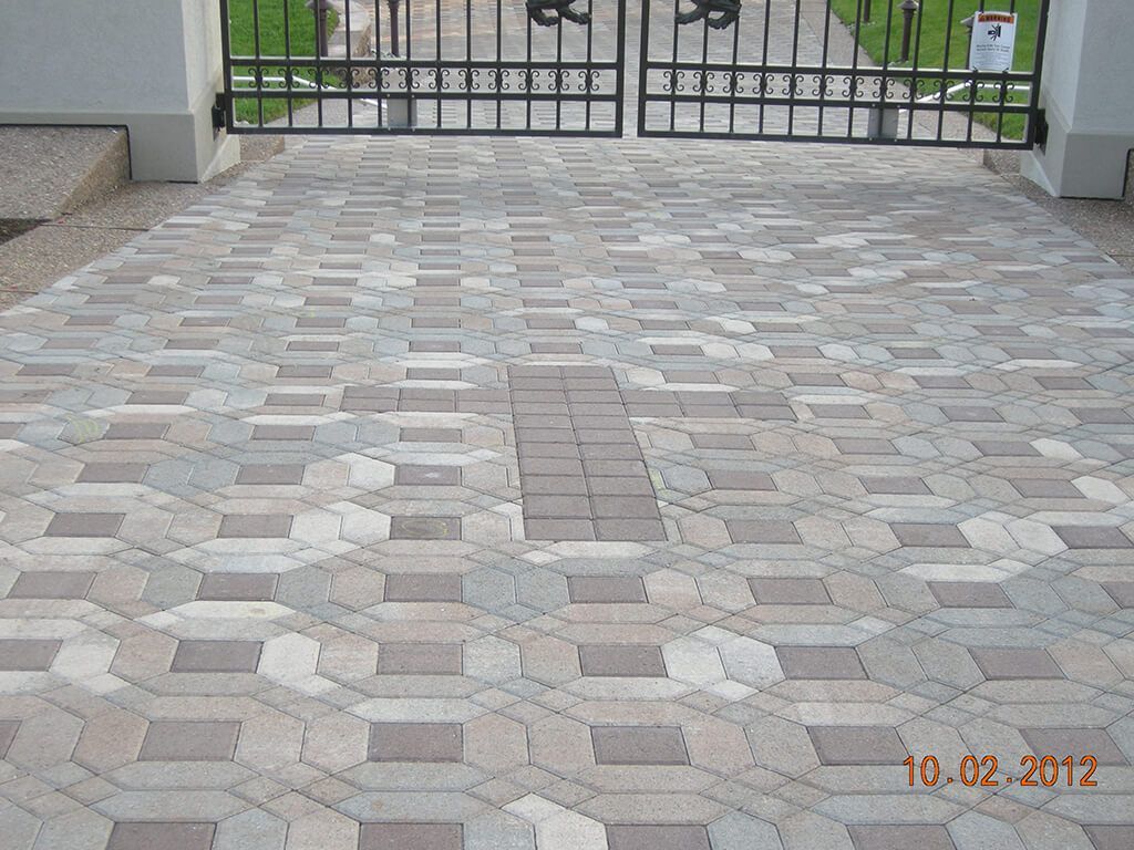 Driveway paved with multi-colored interlocking bricks, leading to a wrought iron gate.