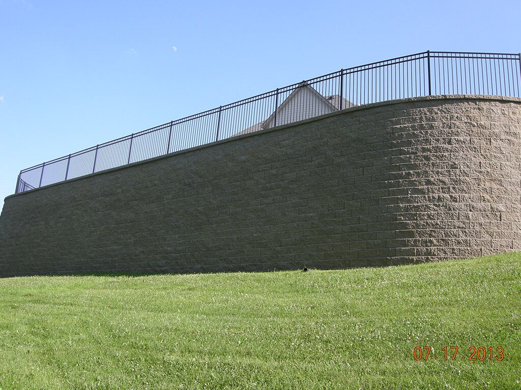 Stone retaining wall with black metal fence on a grassy hill under a blue sky.