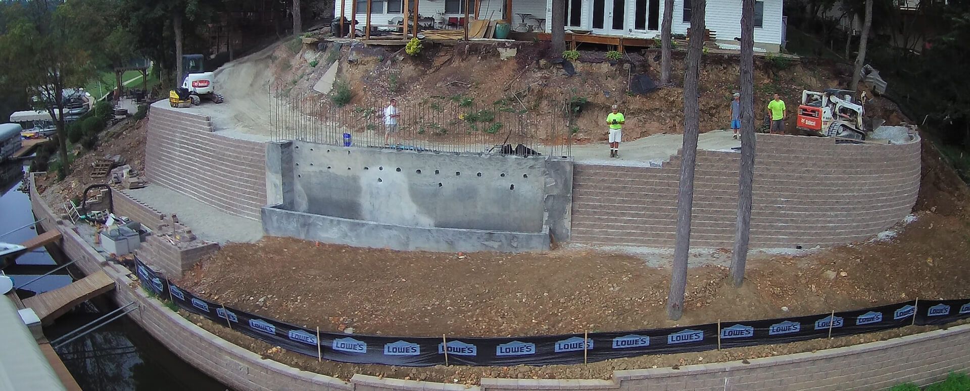 Construction site with retaining walls, concrete work, and a small excavator. Two workers in yellow vests are visible.