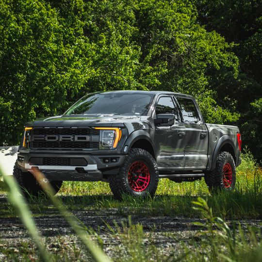 Dark gray Ford Raptor truck with red rims parked on a dirt road surrounded by greenery.