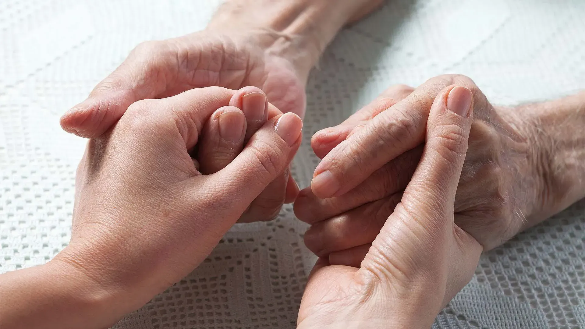 Two pairs of hands, one younger, one older, clasped together on a white surface. Showing comfort and care.