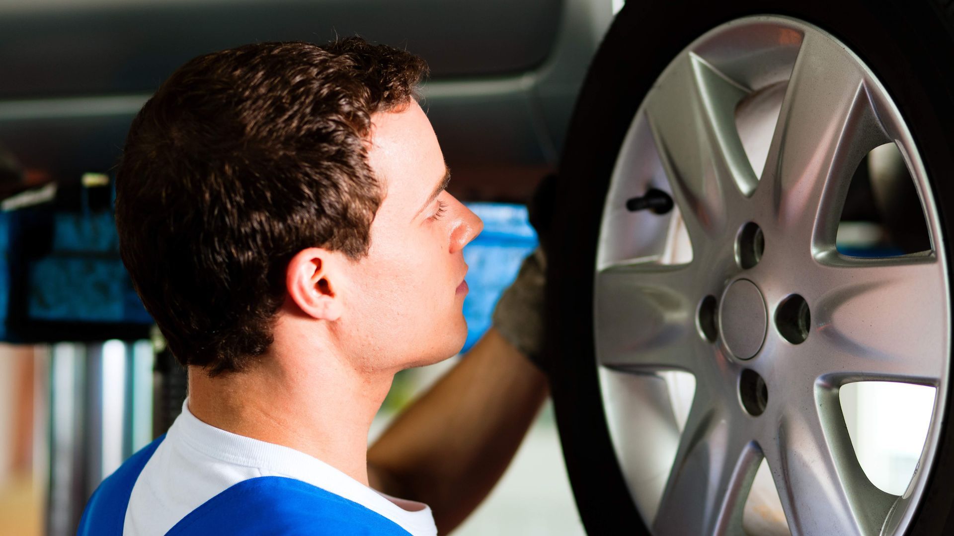 Mechanic checking a car wheel in a garage. He's wearing a blue shirt and looking closely.