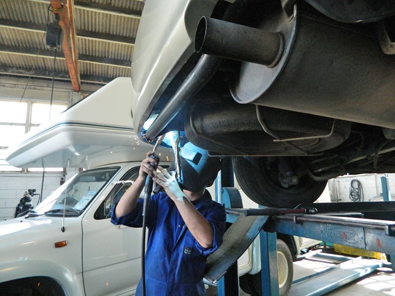 Welder in a blue jumpsuit welding under a white car on a lift in a garage.
