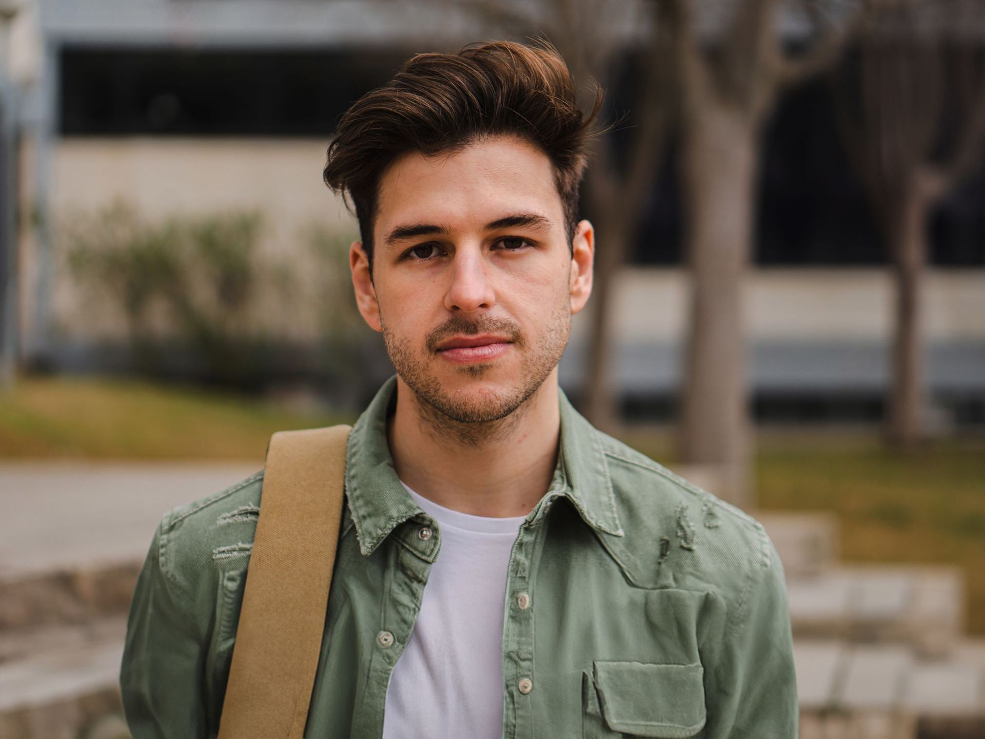 A young man with a beard is wearing a green shirt and carrying a brown bag.