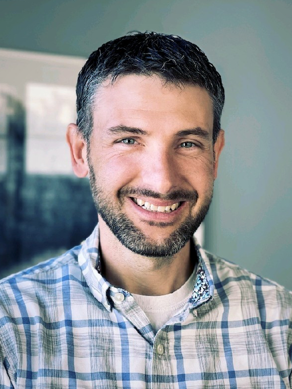 A man with a beard wearing a plaid shirt is smiling for the camera.