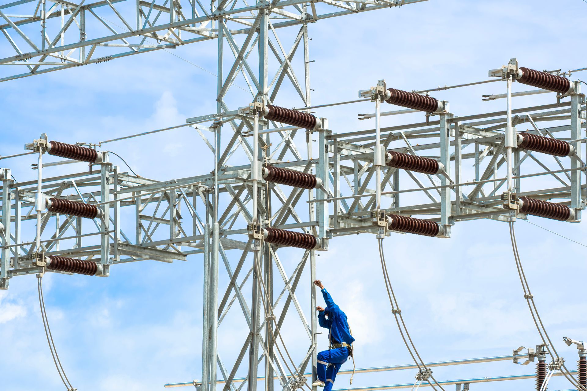 Lineman in blue uniform climbing electrical tower in bright daylight.
