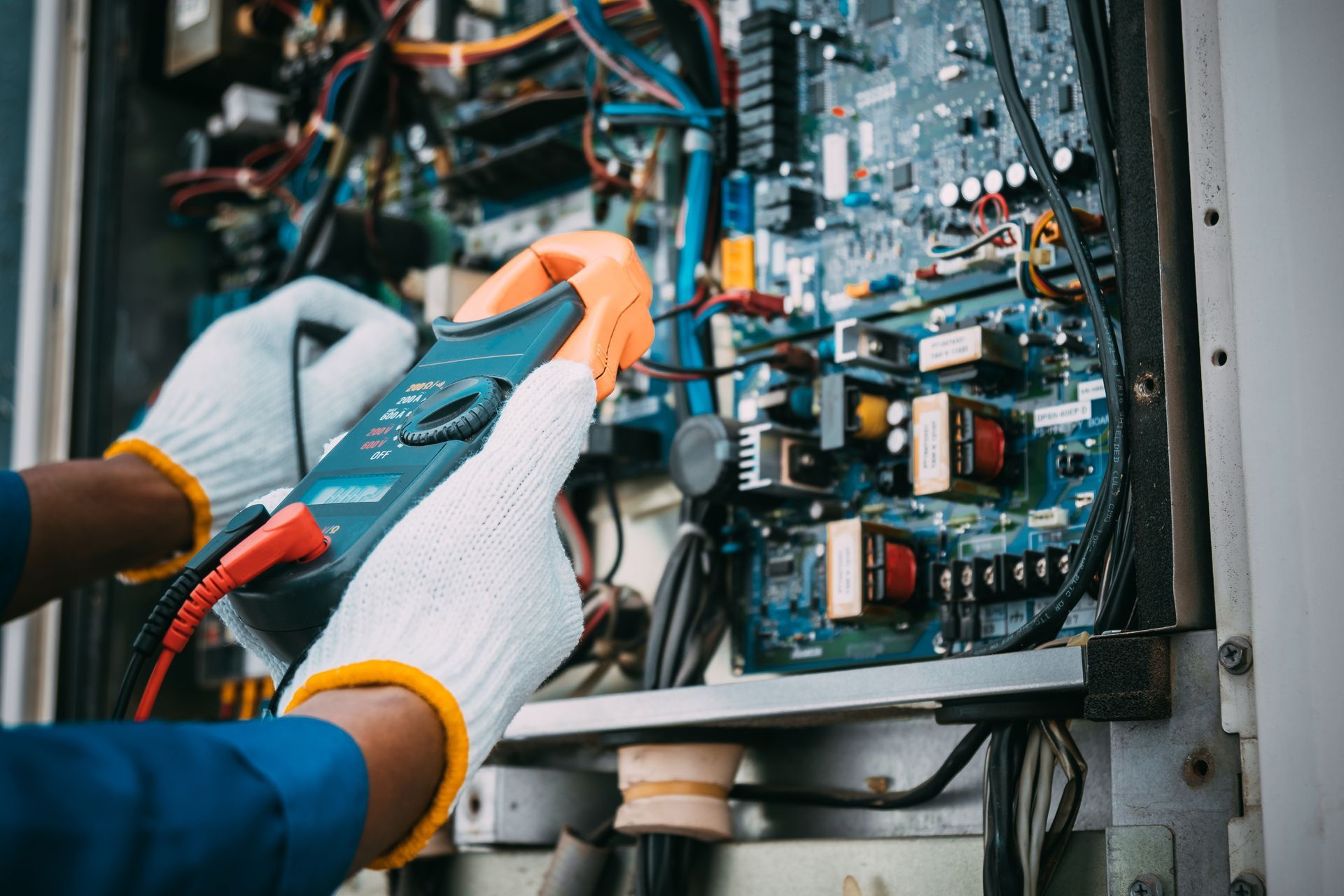 Hands wearing gloves using a multimeter to inspect circuit board inside a machine.