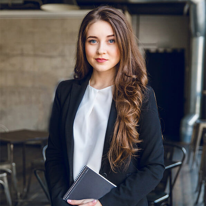Woman in black blazer and white shirt holding notebook, looking at the camera. Indoor setting.