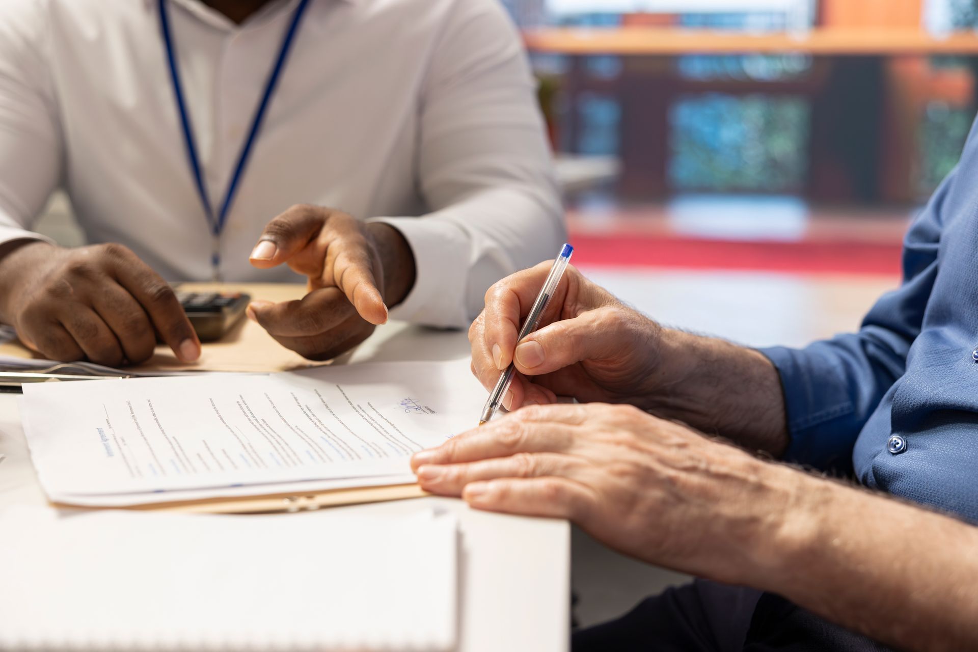 Person signing a document at a table, with another person pointing. White desk, blue shirts.