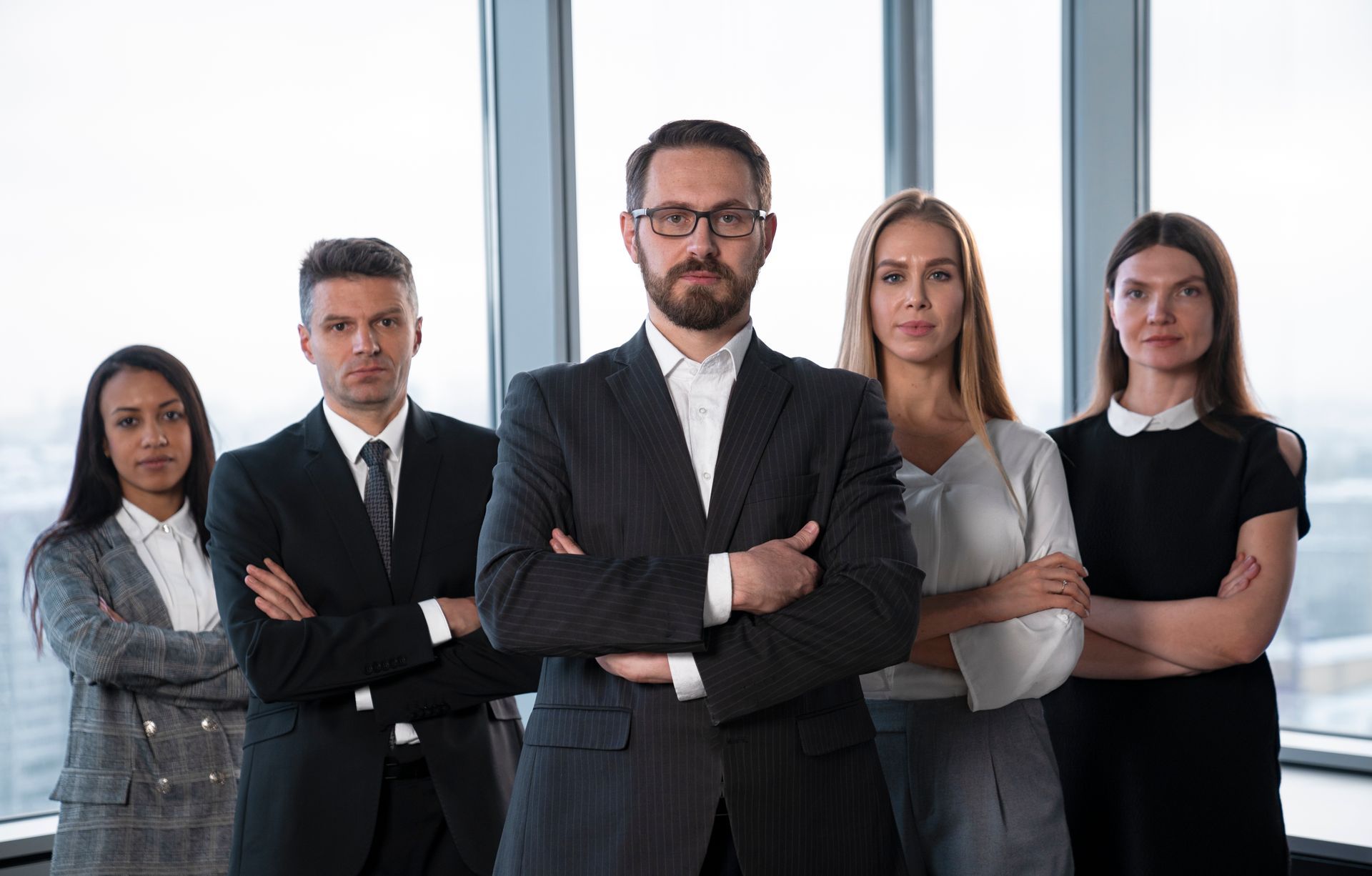 Group of people in business attire standing with arms crossed, looking serious in an office.
