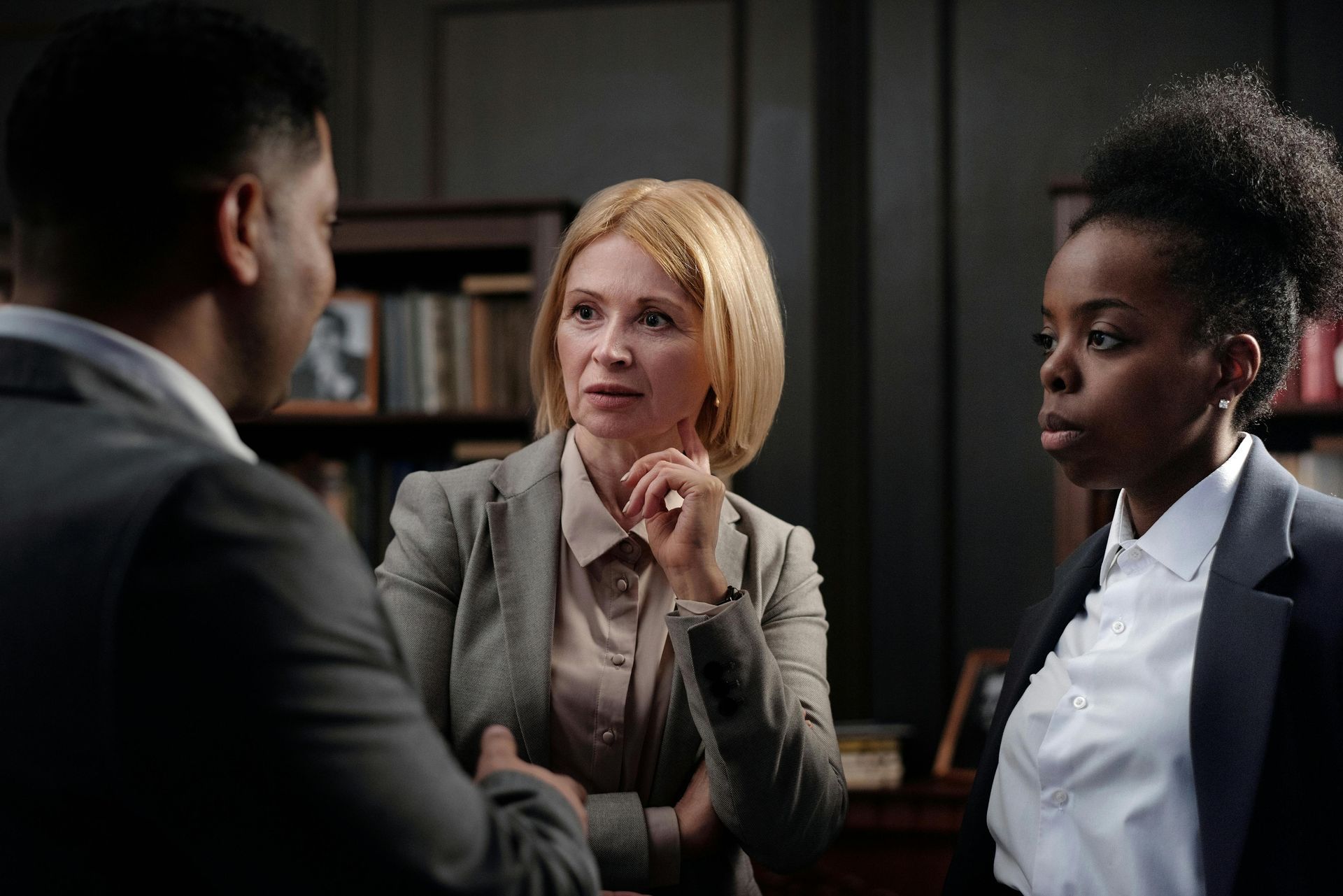 Three people in suits discussing, inside office with bookshelves. Middle person gestures, others listen intently.