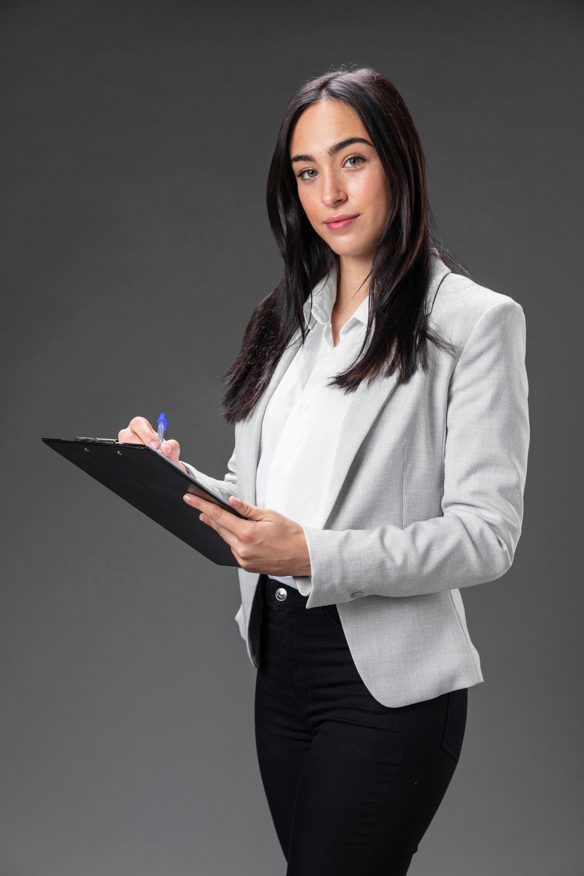 Woman in light blazer writing on clipboard, smiling, against gray background.