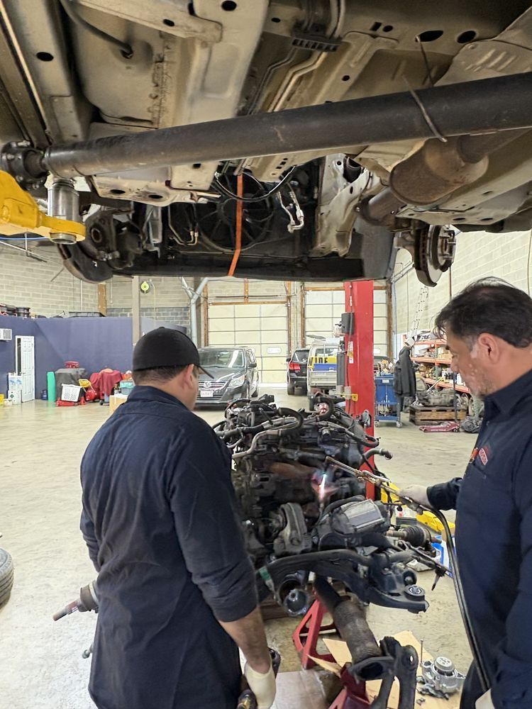 Two mechanics work on a car engine suspended beneath a vehicle lifted on a hydraulic hoist in a repair shop.