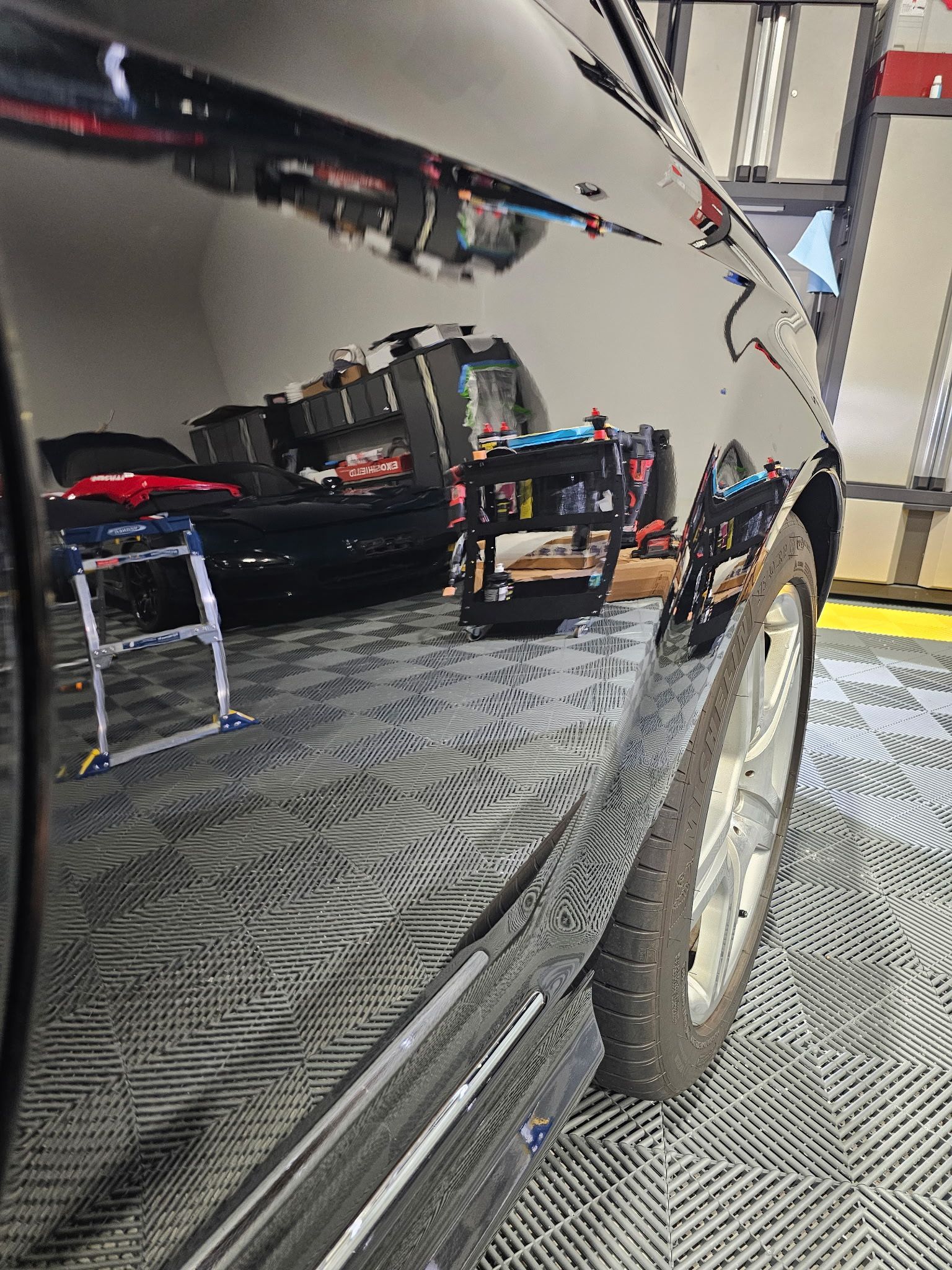 Black car's shiny side reflecting a garage interior with tools and a ladder. Silver wheel visible.