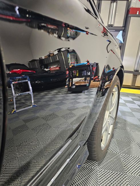 Black car's shiny side reflecting a garage interior with tools and a ladder. Silver wheel visible.