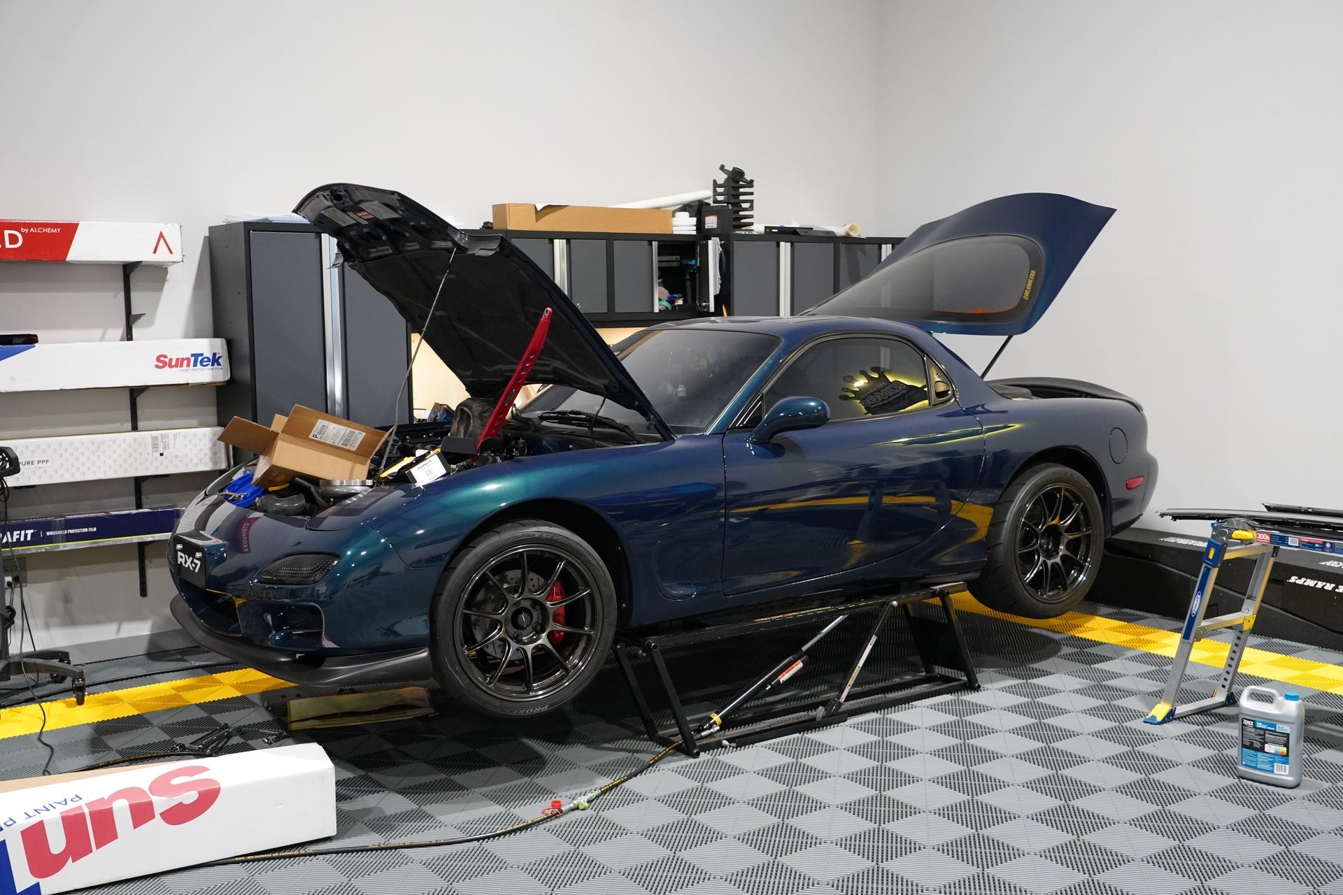 Dark blue Mazda RX-7 with hood and hatch open, on jack stands in a garage.