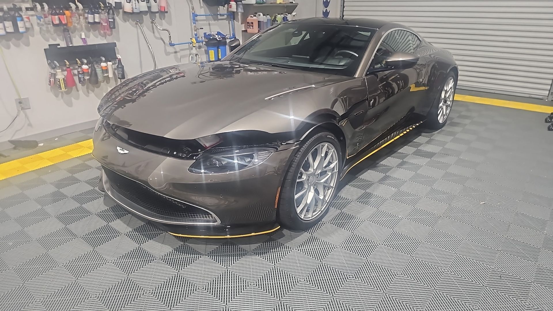 Sleek gray Aston Martin sports car in a garage with shiny chrome wheels, parked on a black and gray tiled floor.
