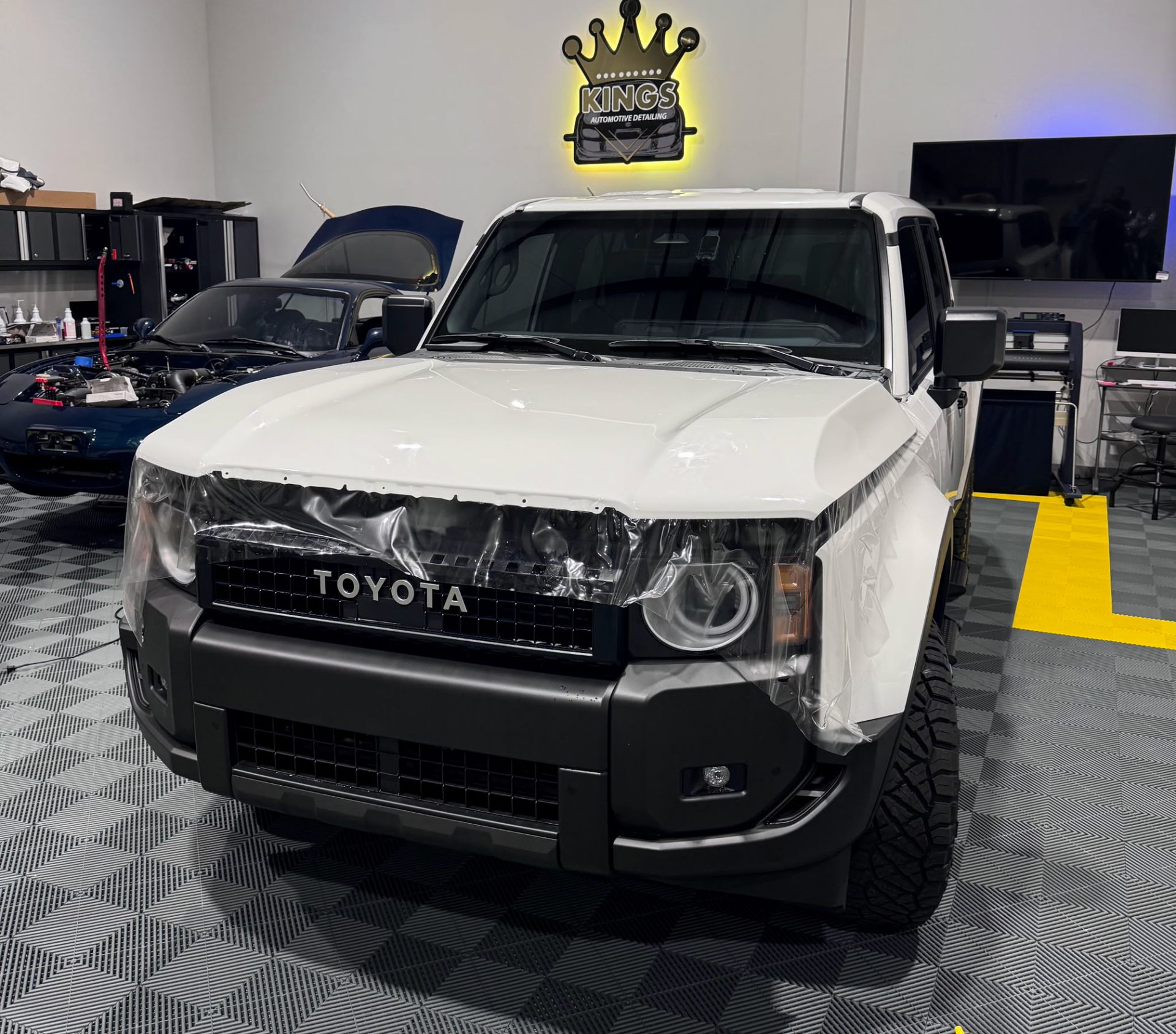 White Toyota truck with protective film on front, in a garage with another vehicle and tools.