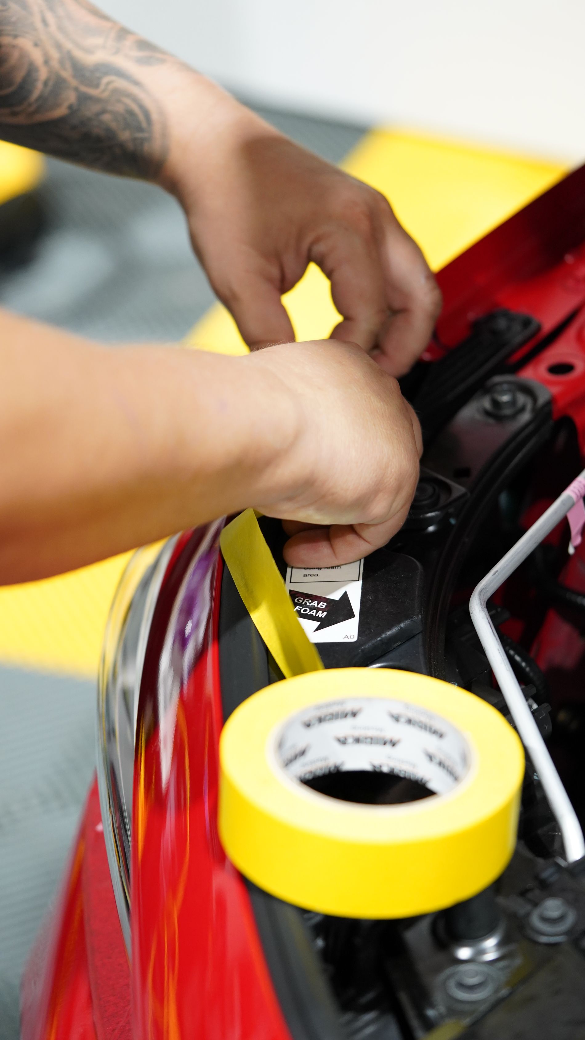 Person taping a red car, using yellow tape in a workshop.