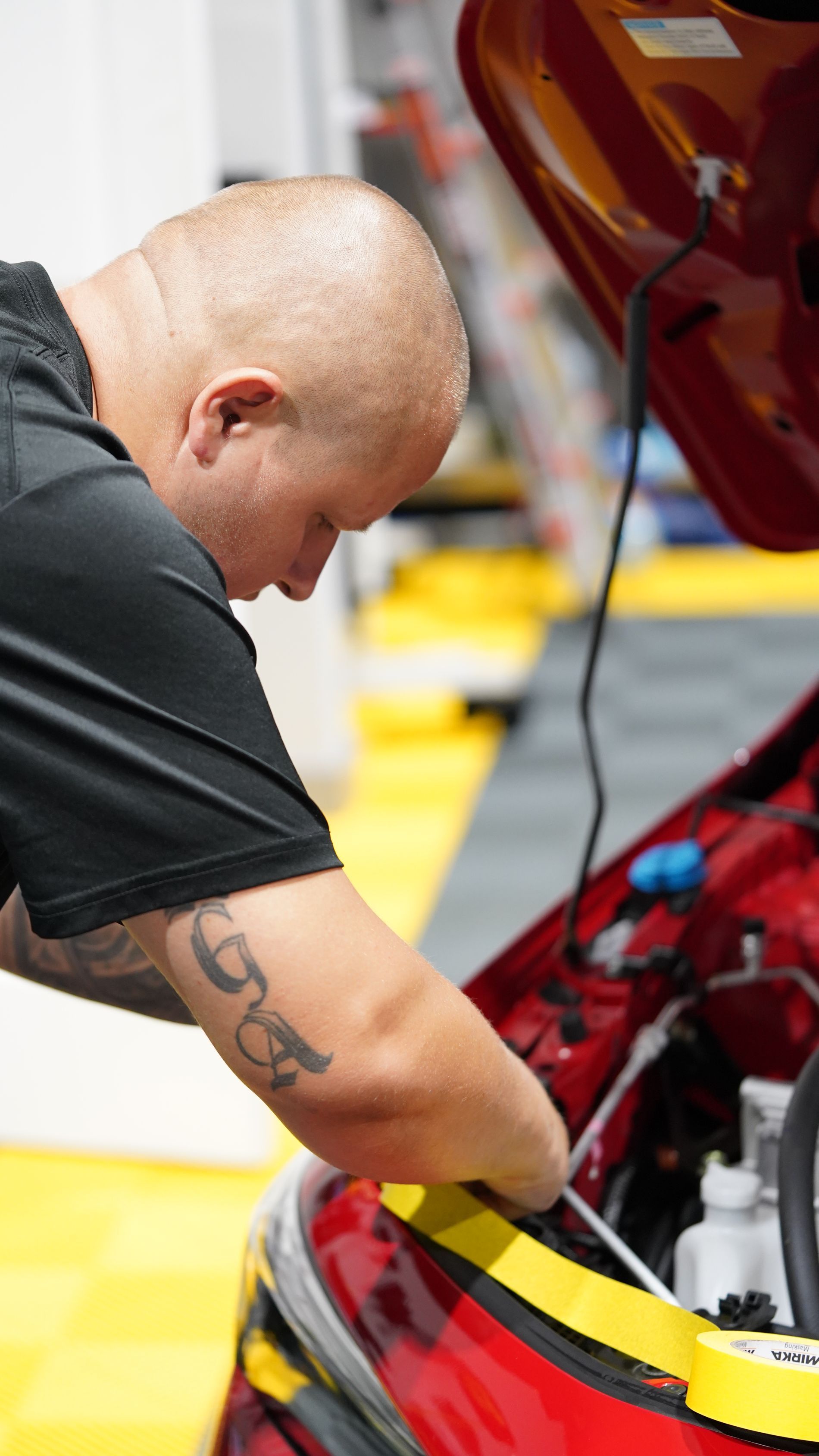 Man with shaved head working under the hood of a red car, yellow floor in background.