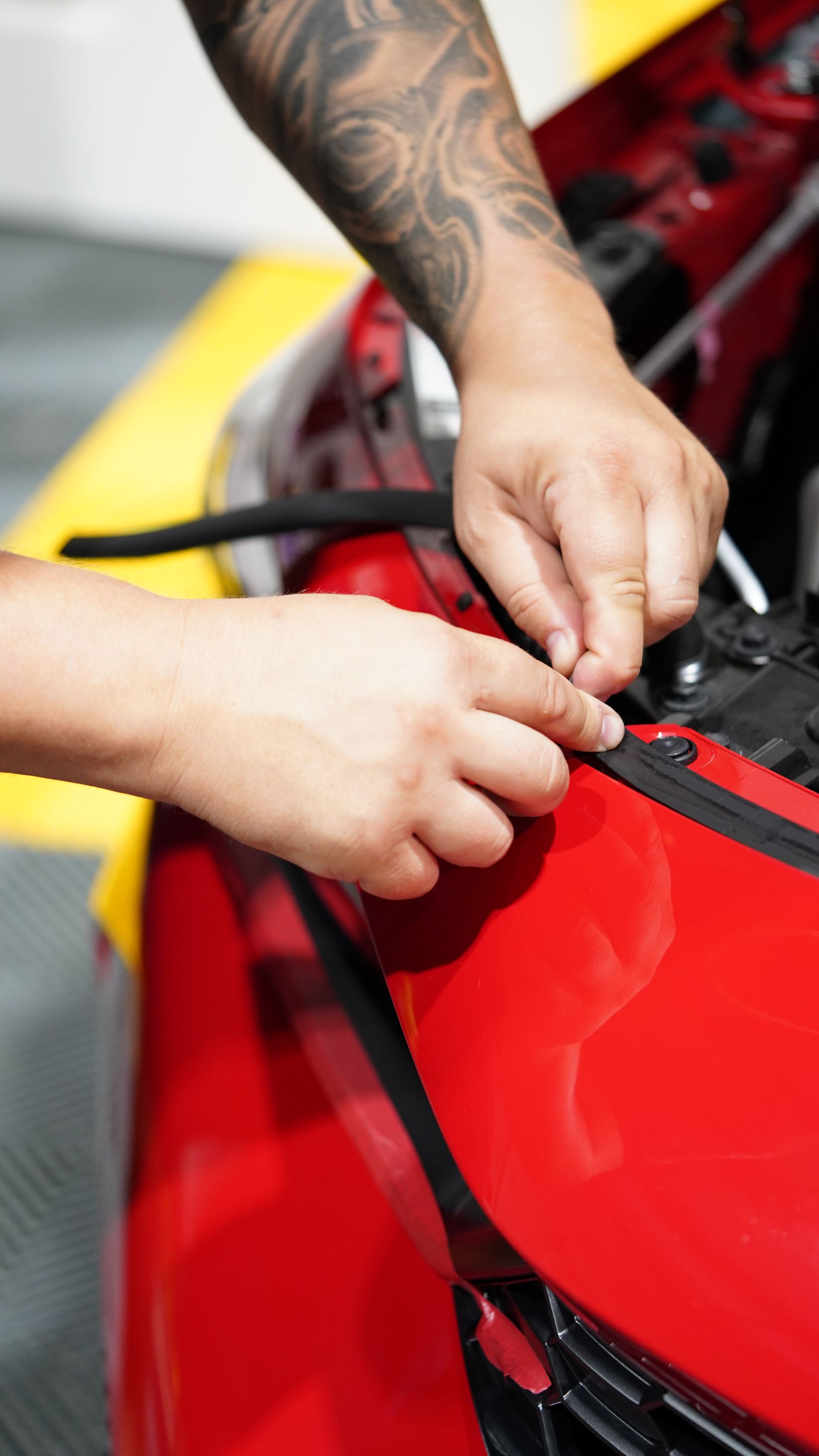 Person with arm tattoos installing black weather stripping on a red car's hood.