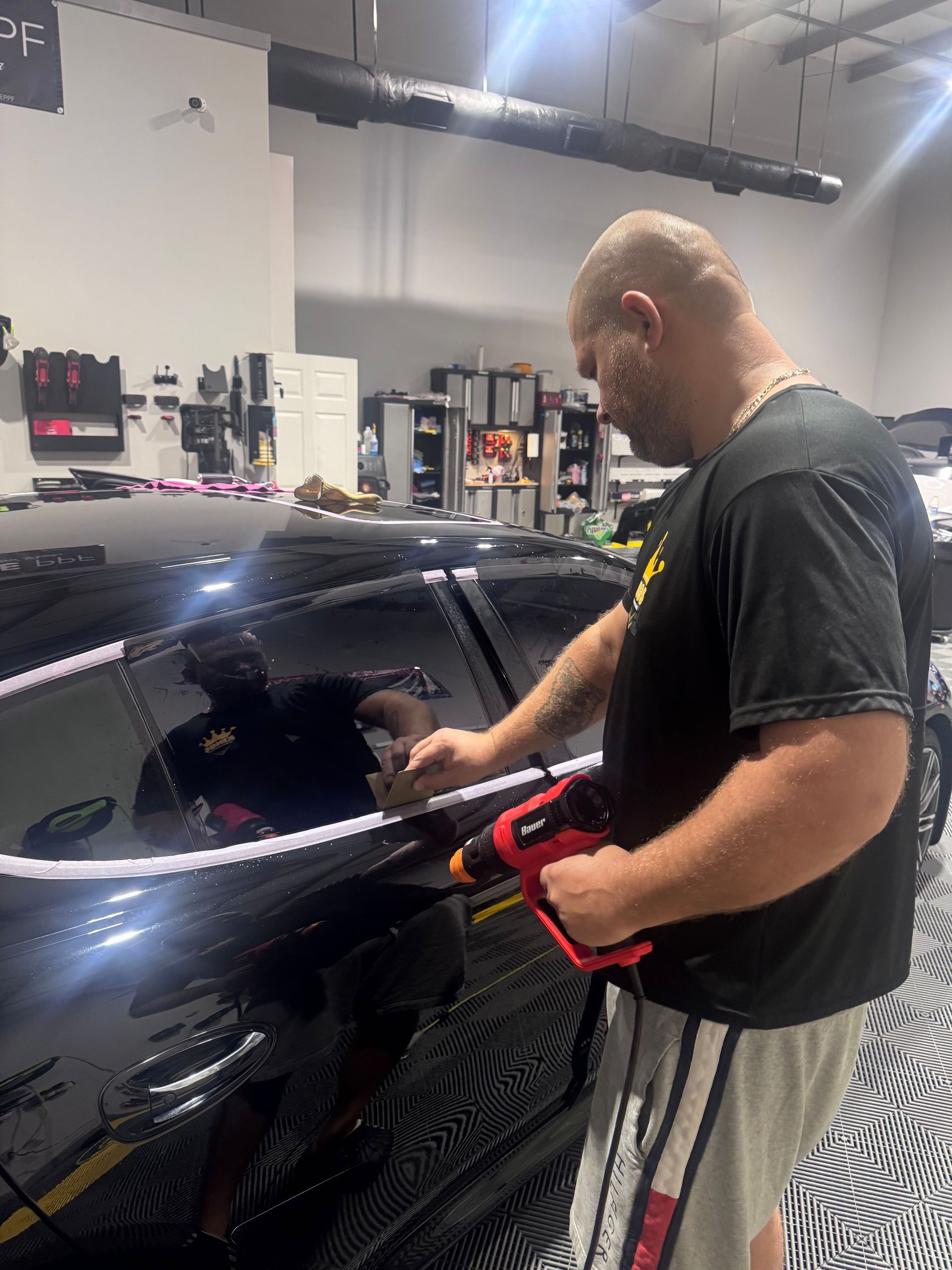 Man applying window tint to a black car with a heat gun in a garage.