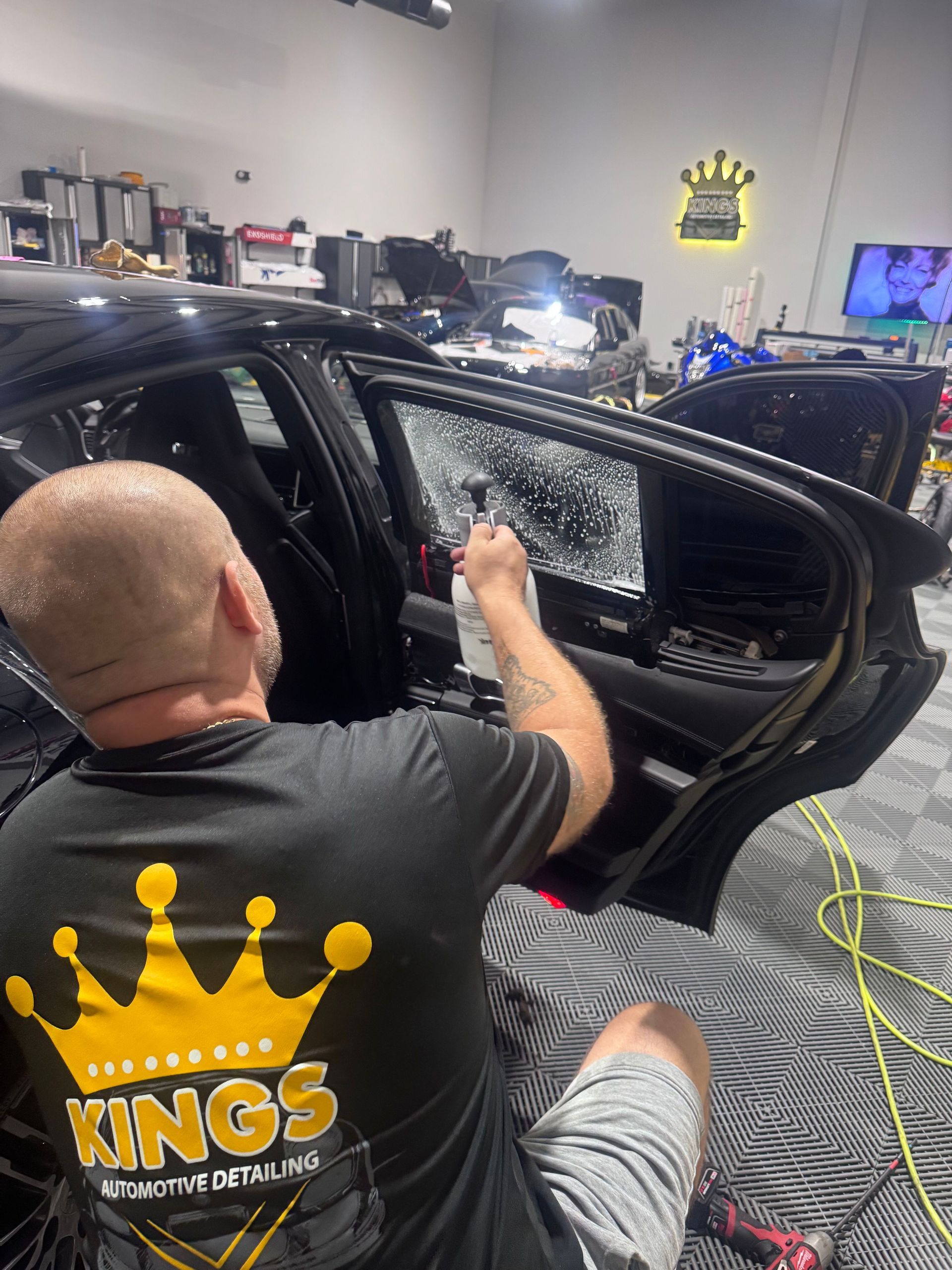 Man applying tint to a car window in a detailing shop. Gold crown logo visible on shirt and wall.